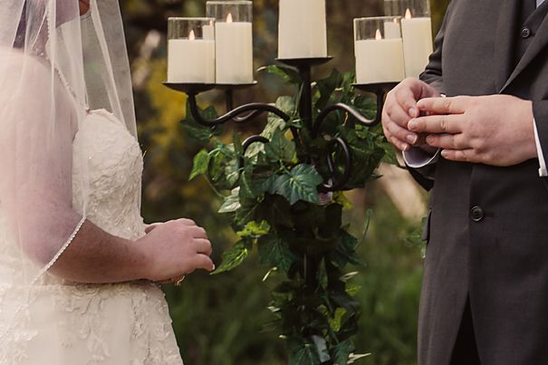 A bride and groom hold hands during an outdoor wedding ceremony with greenery in the background.