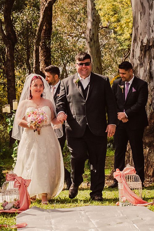 Wedding party poses together outdoors among trees with pink decorative accents visible in the foreground.