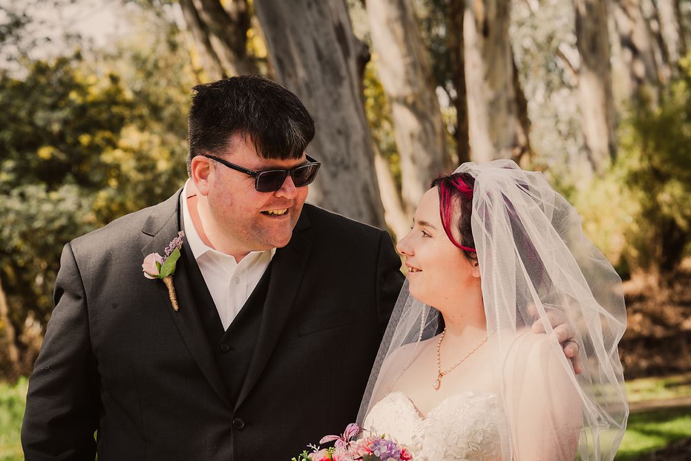 A romantic wedding couple shares tender moments in a garden setting with natural light filtering through trees.