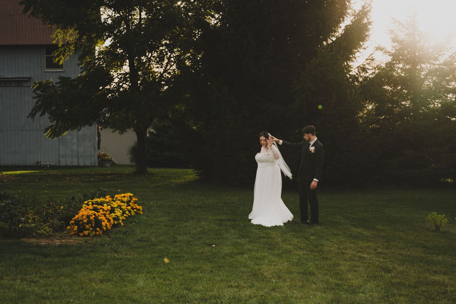 A couple in wedding attire poses in a sunlit garden during golden hour as their photographer captures the moment.