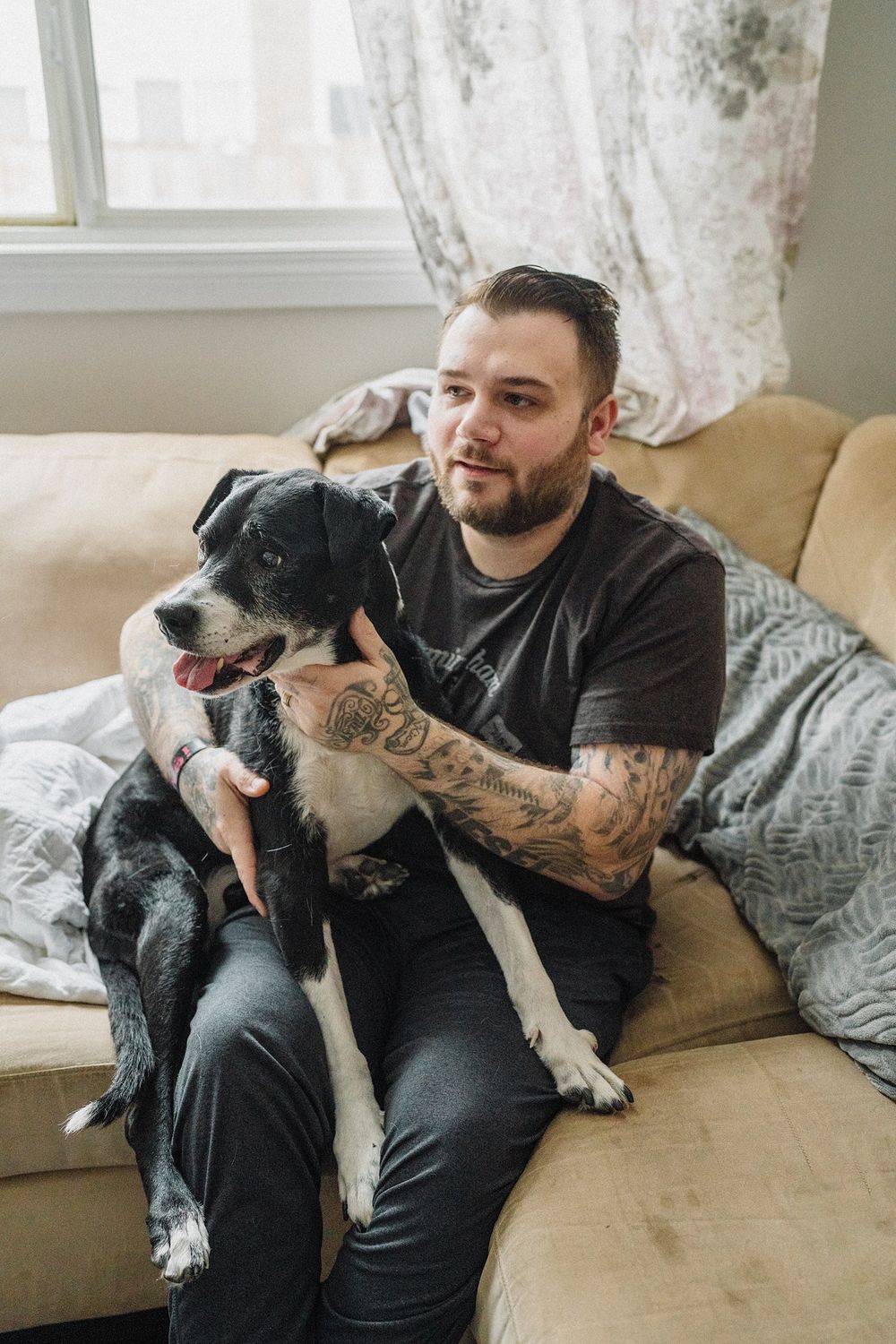 Tattooed man sitting on beige couch holding his black and white dog by window.