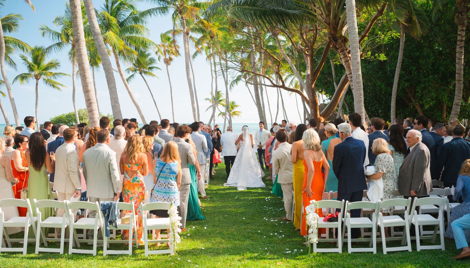 Wedding guests gather under palm trees at a tropical beach ceremony with white chairs and orange accents.