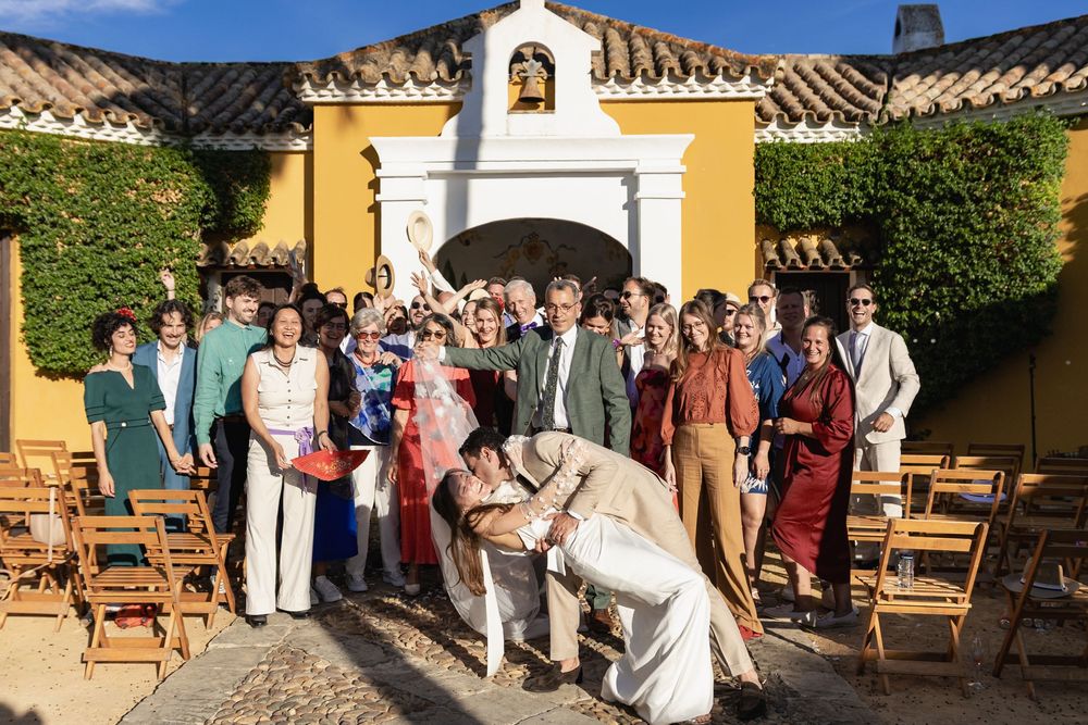 Wedding guests gather in a rustic courtyard with yellow walls and wooden benches during an outdoor celebration.