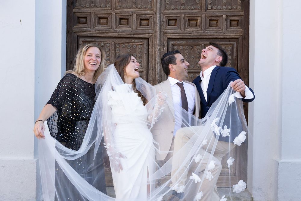 Wedding party laughs joyfully on church steps while holding flowing white veil in the breeze.