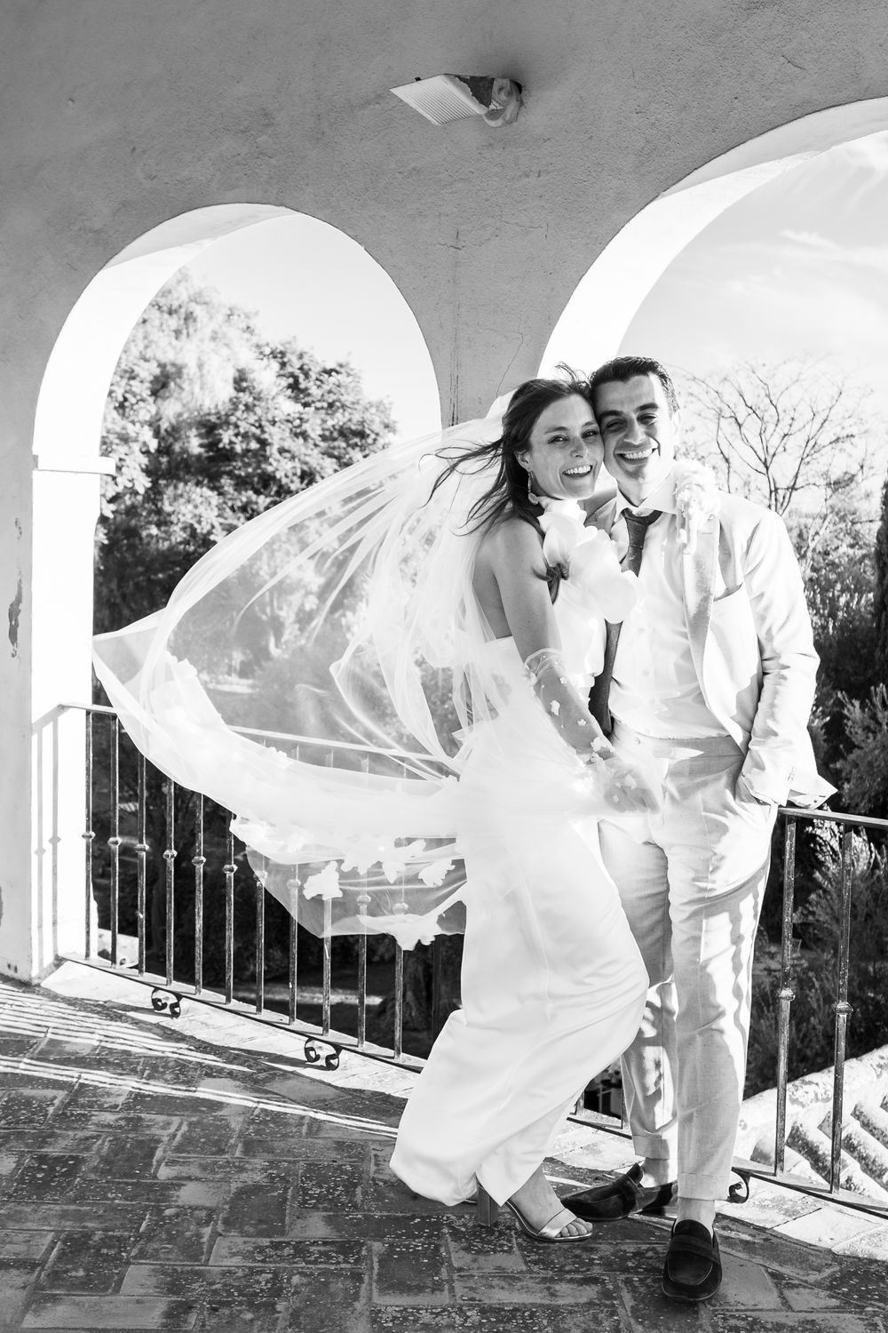A flowing white wedding veil billows dramatically against an arched architectural backdrop in black and white.