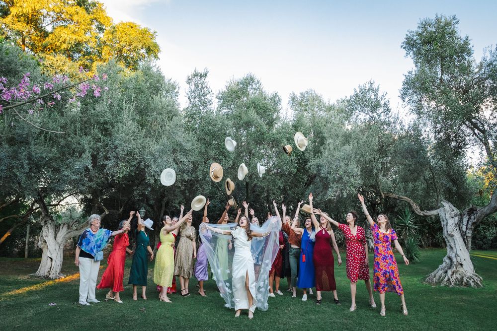A group of wedding guests in colorful dresses throw their hats in the air beneath olive trees at sunset.