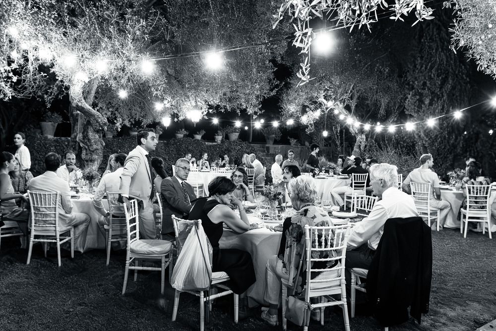String lights illuminate an outdoor evening wedding reception with guests seated at tables under trees.