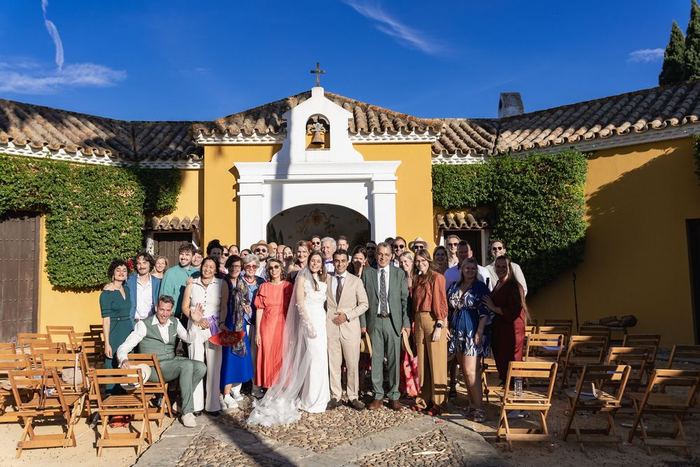 Wedding guests gather in a rustic courtyard with wooden chairs and yellow stucco walls under bright blue skies.