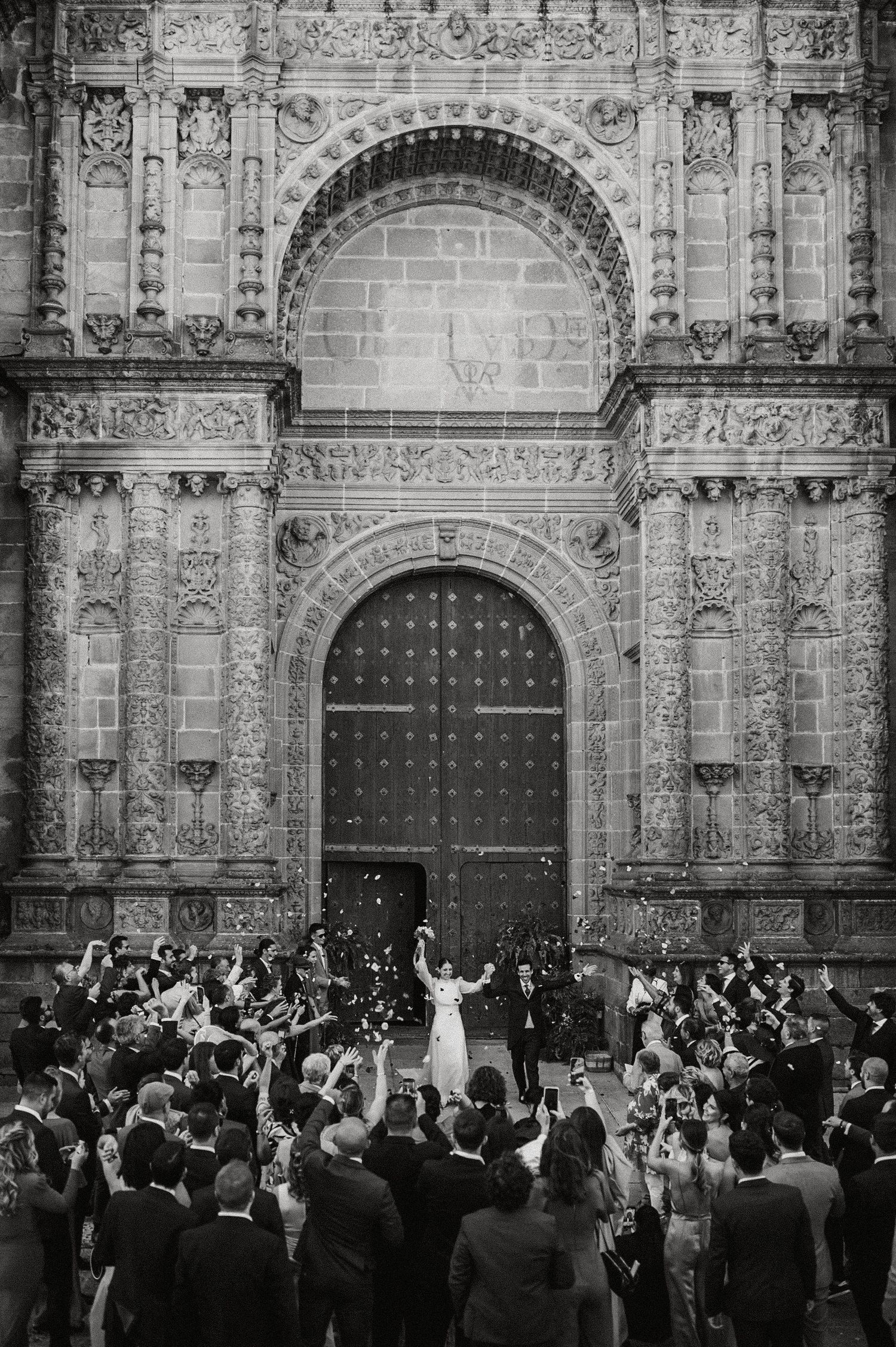 A crowd gathers for a celebration in front of an ornate cathedral with detailed stone carvings and large wooden doors.