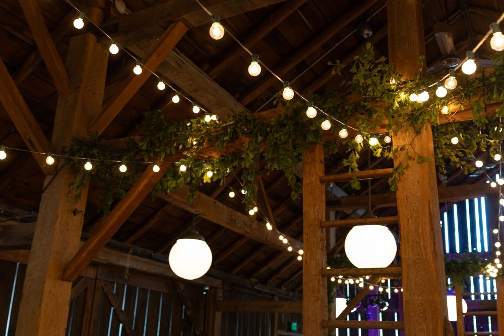 String lights and round white lanterns illuminate a rustic wooden barn interior with exposed beams and greenery.