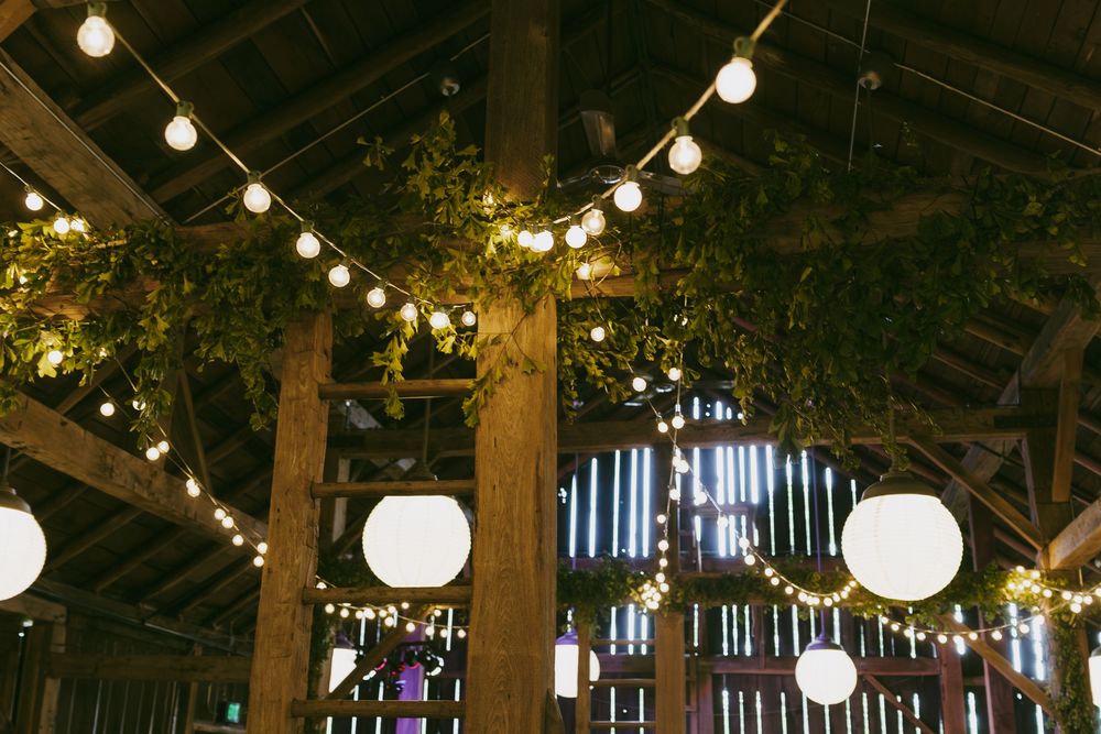 String lights and paper lanterns illuminate a rustic barn interior creating a warm, romantic atmosphere for an event.