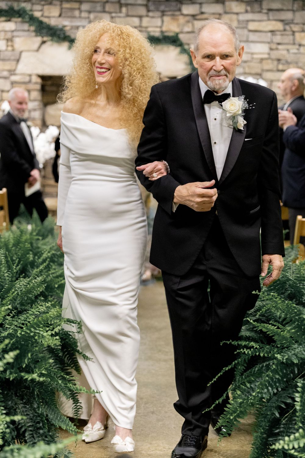 A bride in a white form-fitting dress and voluminous curly hair walks arm-in-arm with a gentleman in a black tuxedo.