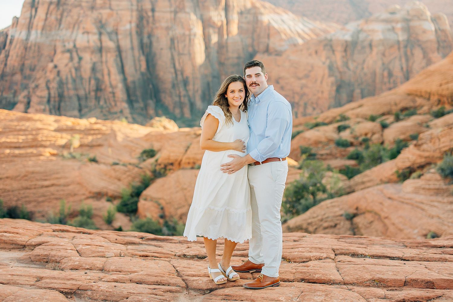 A couple poses together against stunning red rock formations in a desert landscape at sunset.