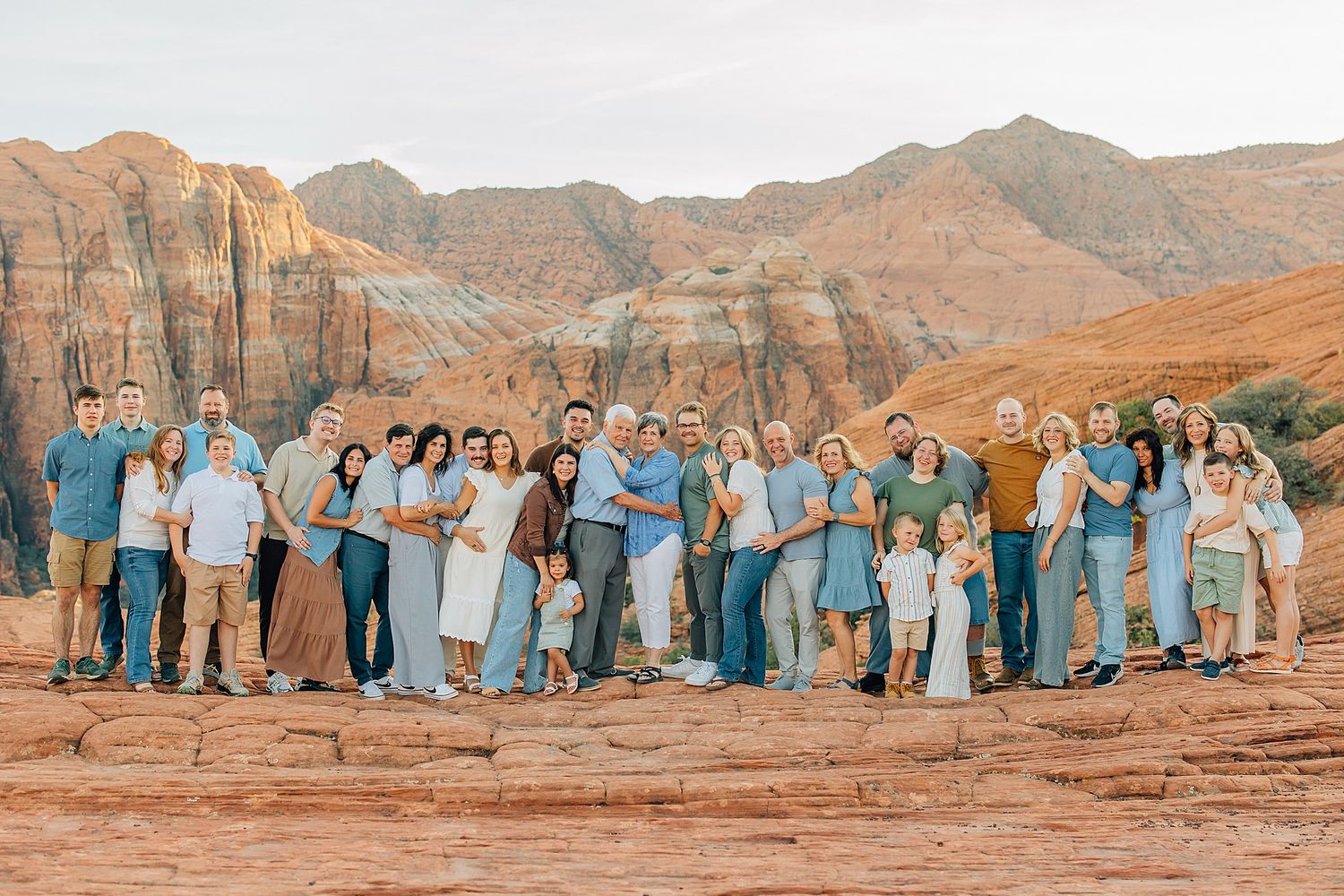 Large family group in coordinated blue and white outfits posing against stunning red rock formations at sunset.