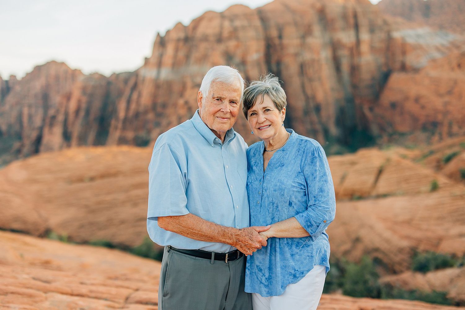 Elderly couple embraces while standing in front of majestic red rock cliffs in desert setting.