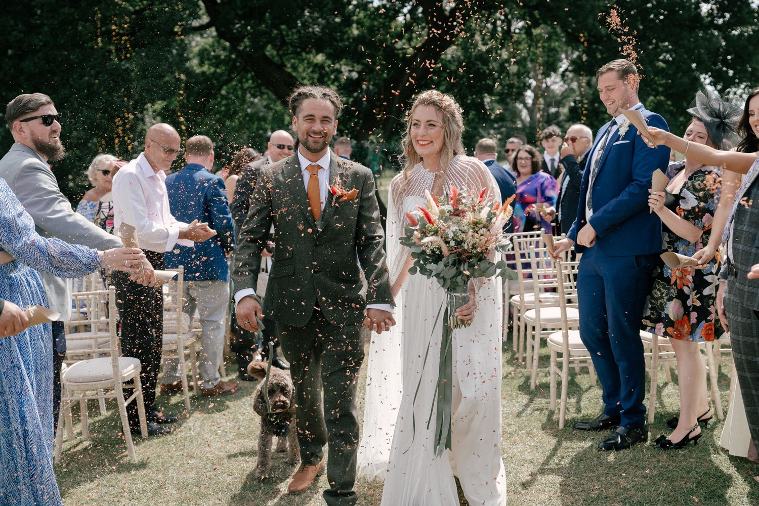 Wedding guests toss confetti and celebrate as a couple walks down an outdoor aisle surrounded by trees and chairs.