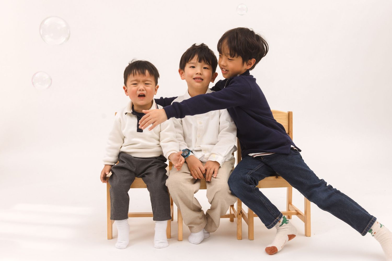 Three children sit and interact playfully together on wooden chairs against a white studio backdrop.