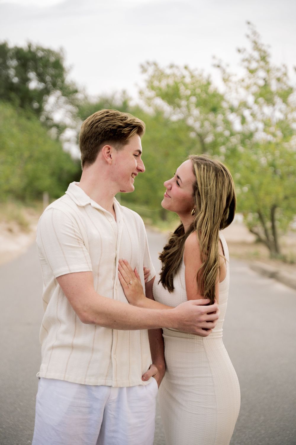 A romantic couple in white outfits sharing an intimate moment on a tree-lined path.
