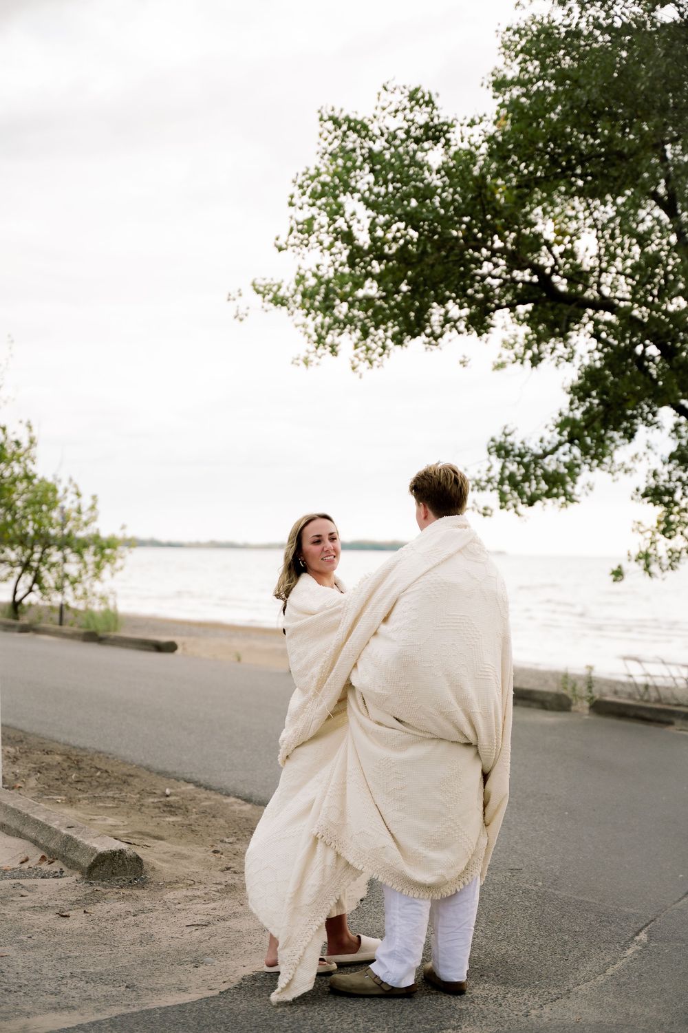 Couple wrapped in a cream blanket sits together on a beachside path during a romantic moment at sunset.