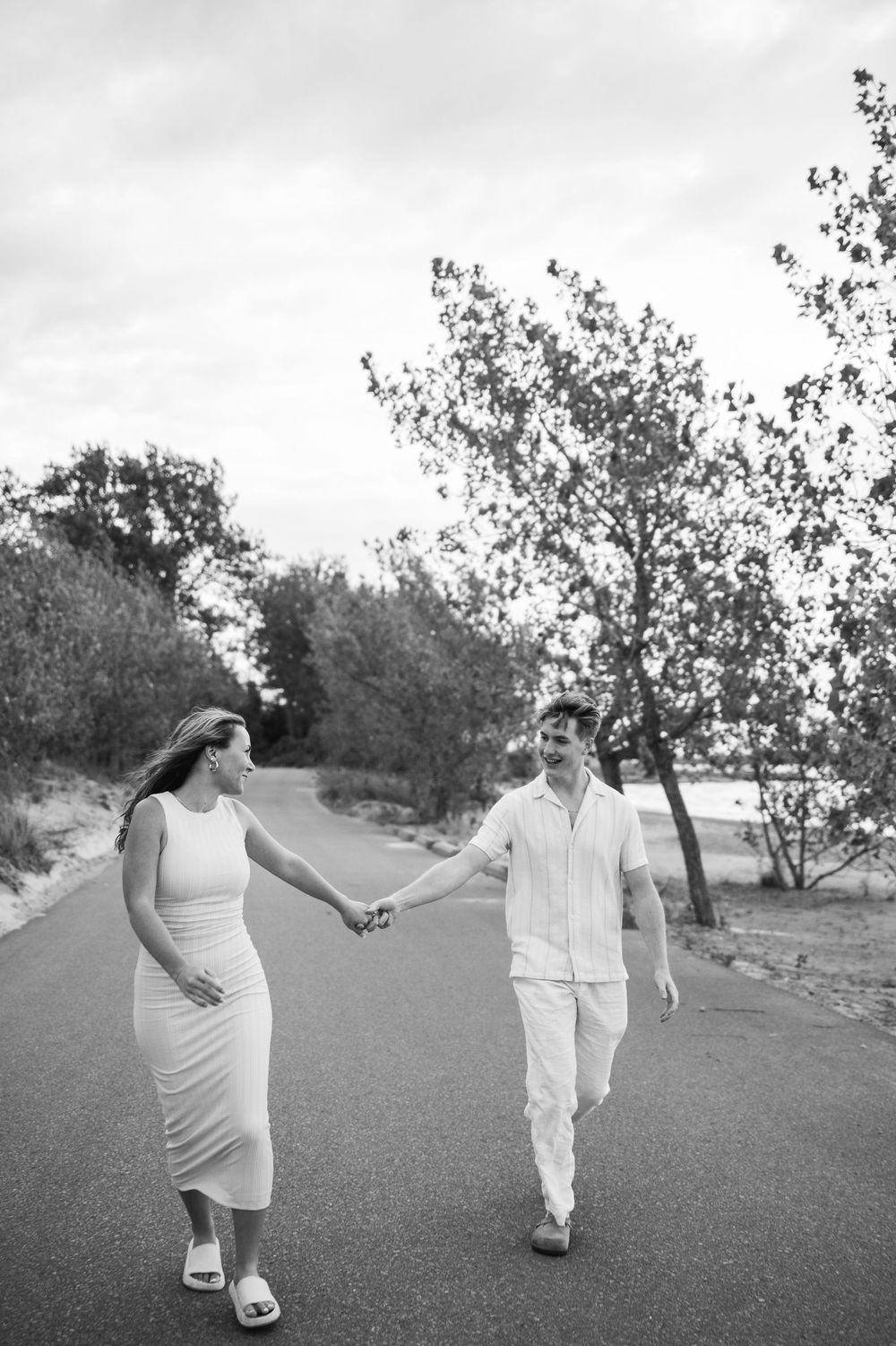 Playful couple in white outfits dances and spins on a grassy hillside under cloudy skies.
