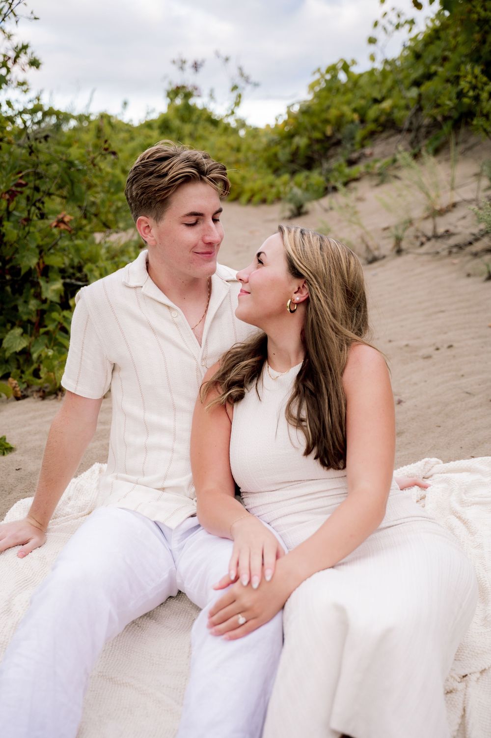 A couple in white clothing sitting together on sandy dunes surrounded by greenery.