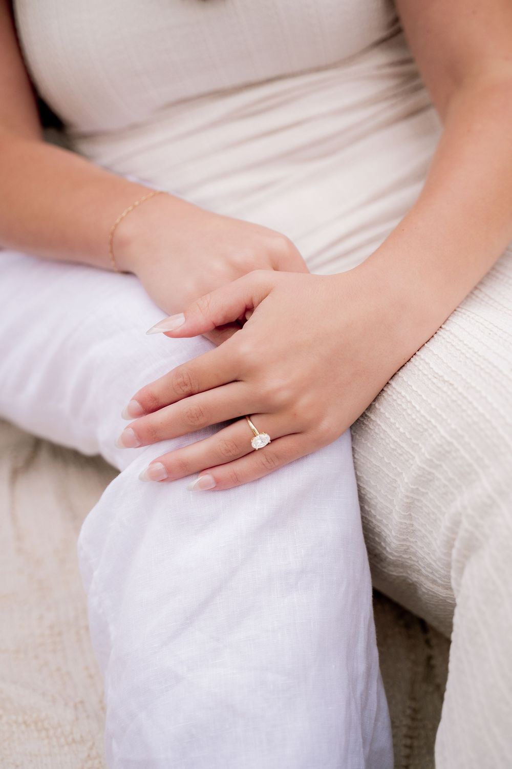 Close-up of hands resting on white fabric showing a delicate engagement ring.