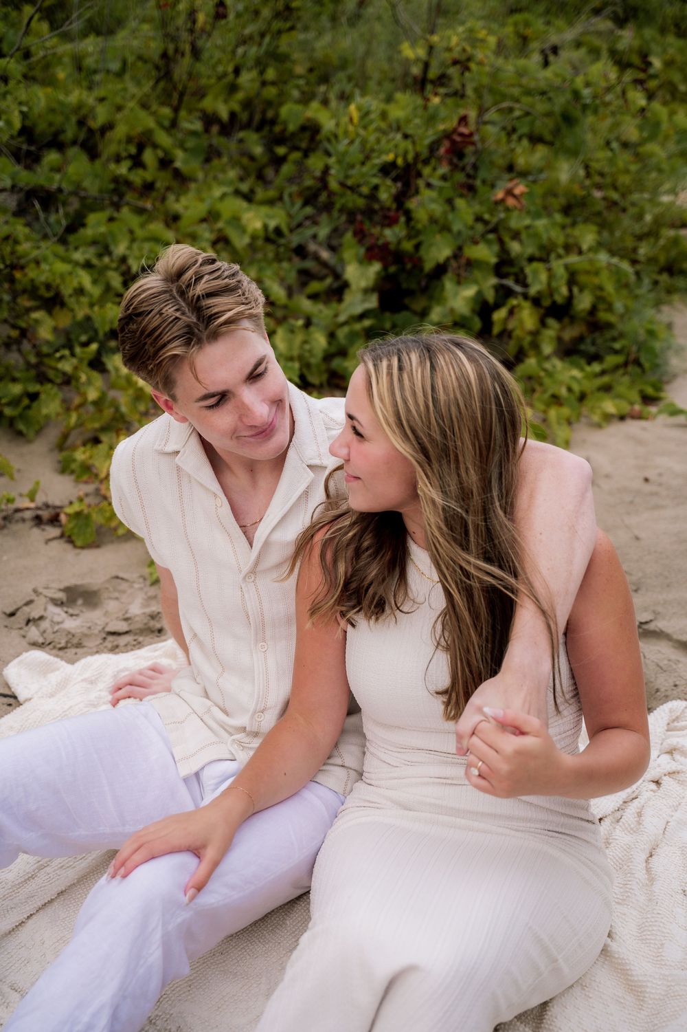 A romantic couple in white clothing lying together on a blanket surrounded by greenery.