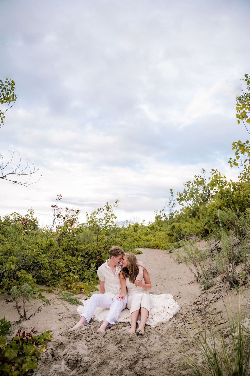 A romantic beach engagement photo shows a couple sitting together on sandy dunes surrounded by coastal vegetation.