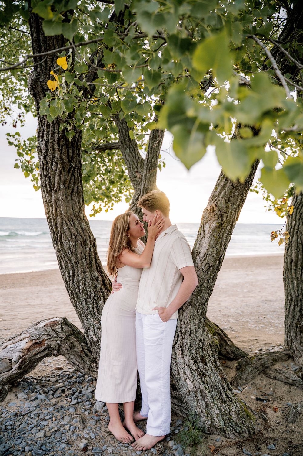 A romantic couple in white outfits embrace under a tree by the beach during a sunset photo session.
