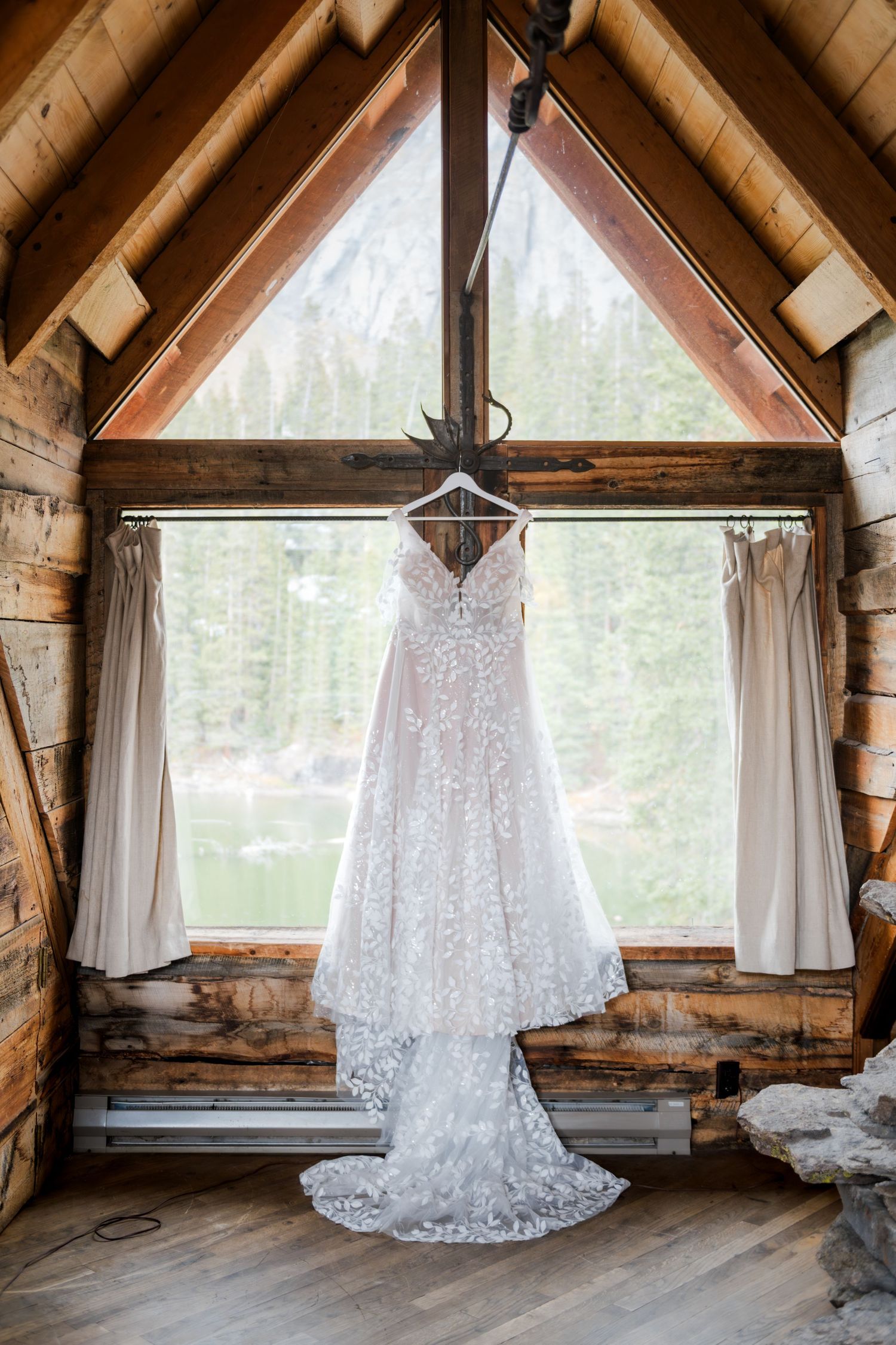 A white lace wedding dress hangs in a rustic wooden attic with natural light streaming through triangular windows.
