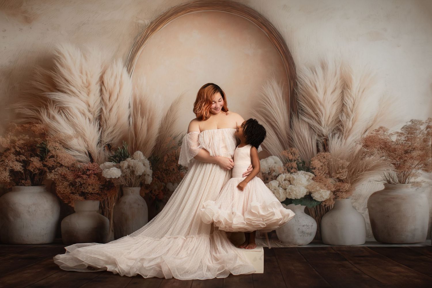 Two people in flowing white dresses pose together against a backdrop of pampas grass and ceramic vases.