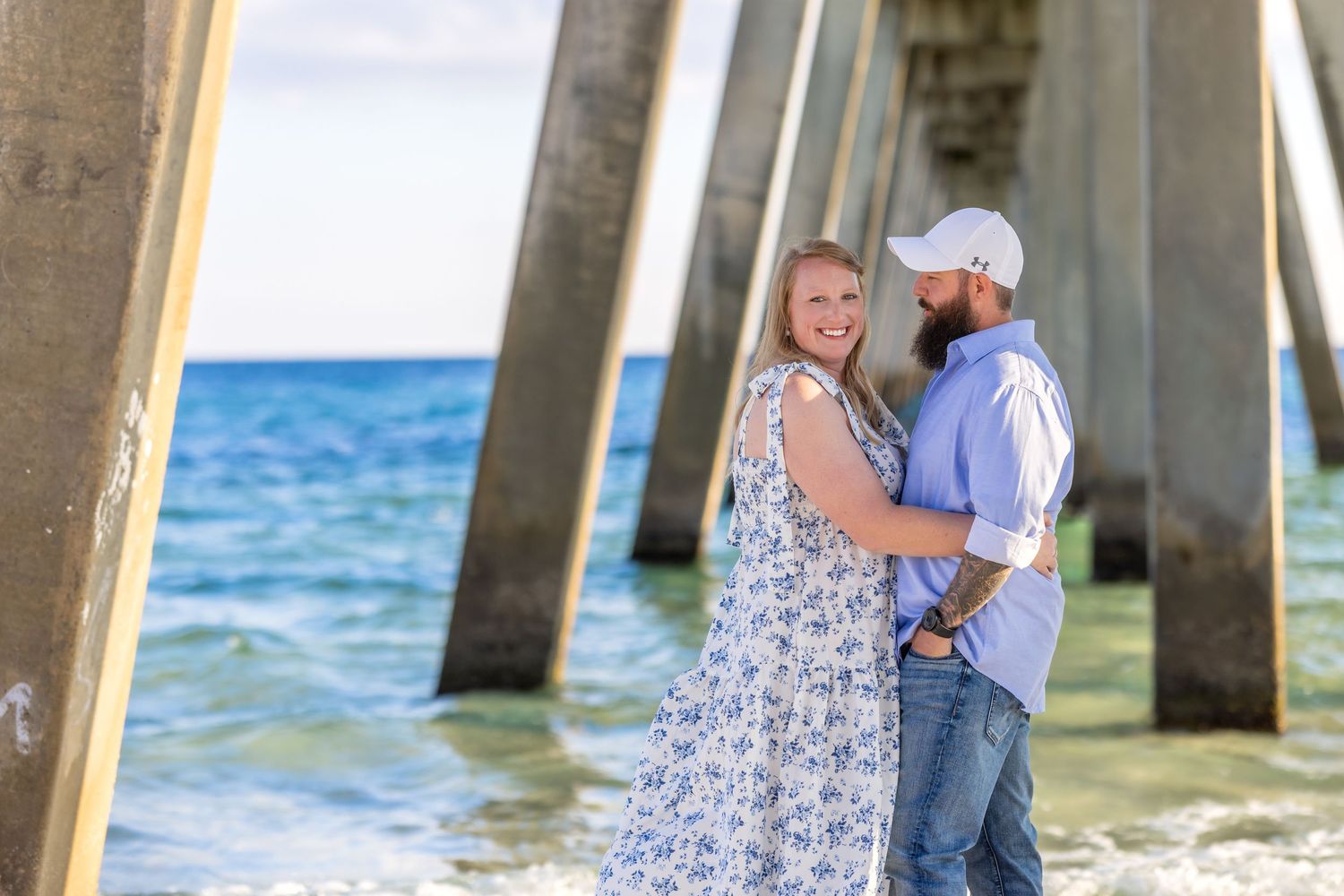 A couple embraces on a sandy beach at sunset while ocean waves roll onto the shore in the background.
