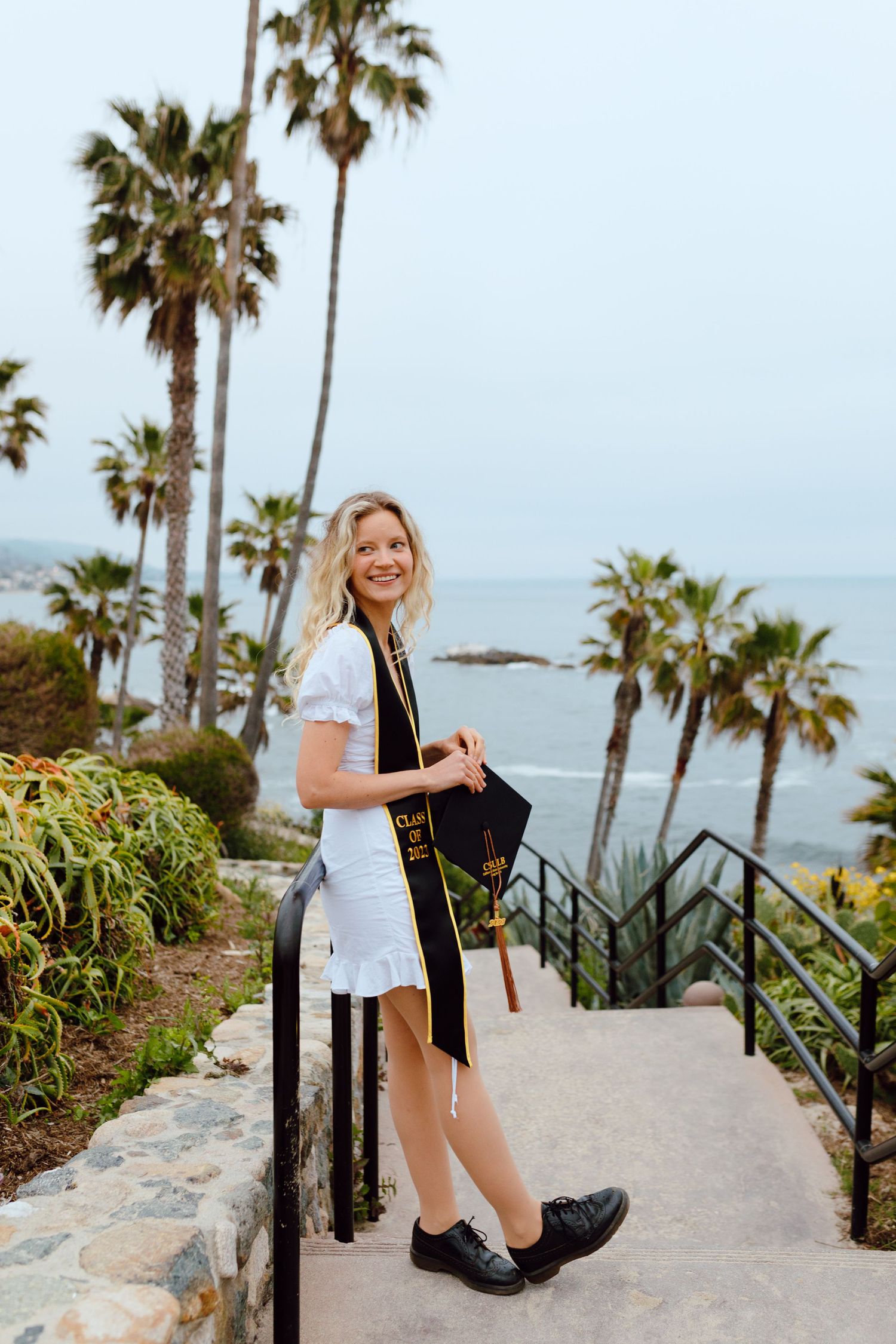 A college graduate in a white dress takes her graduation portraits at a beach.