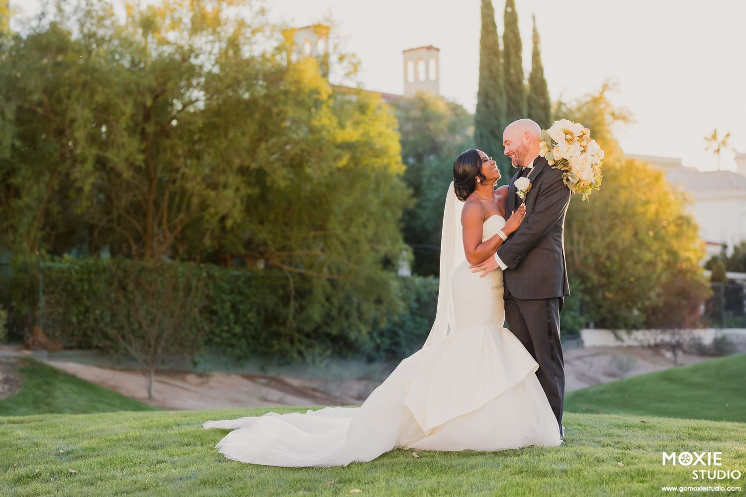 A romantic wedding couple embraces on a lush green lawn at sunset with cypress trees and a building in the background.