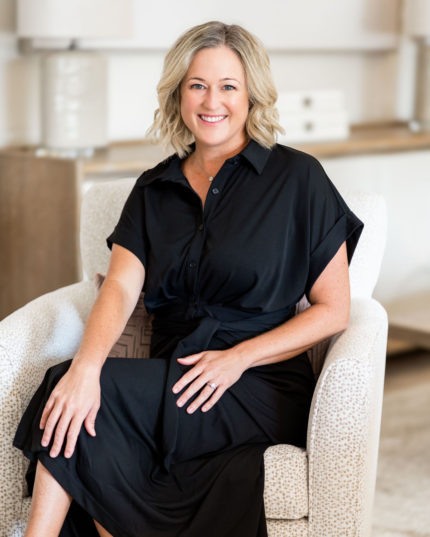 Stylish woman in black dress sitting on white textured chair in bright modern interior.