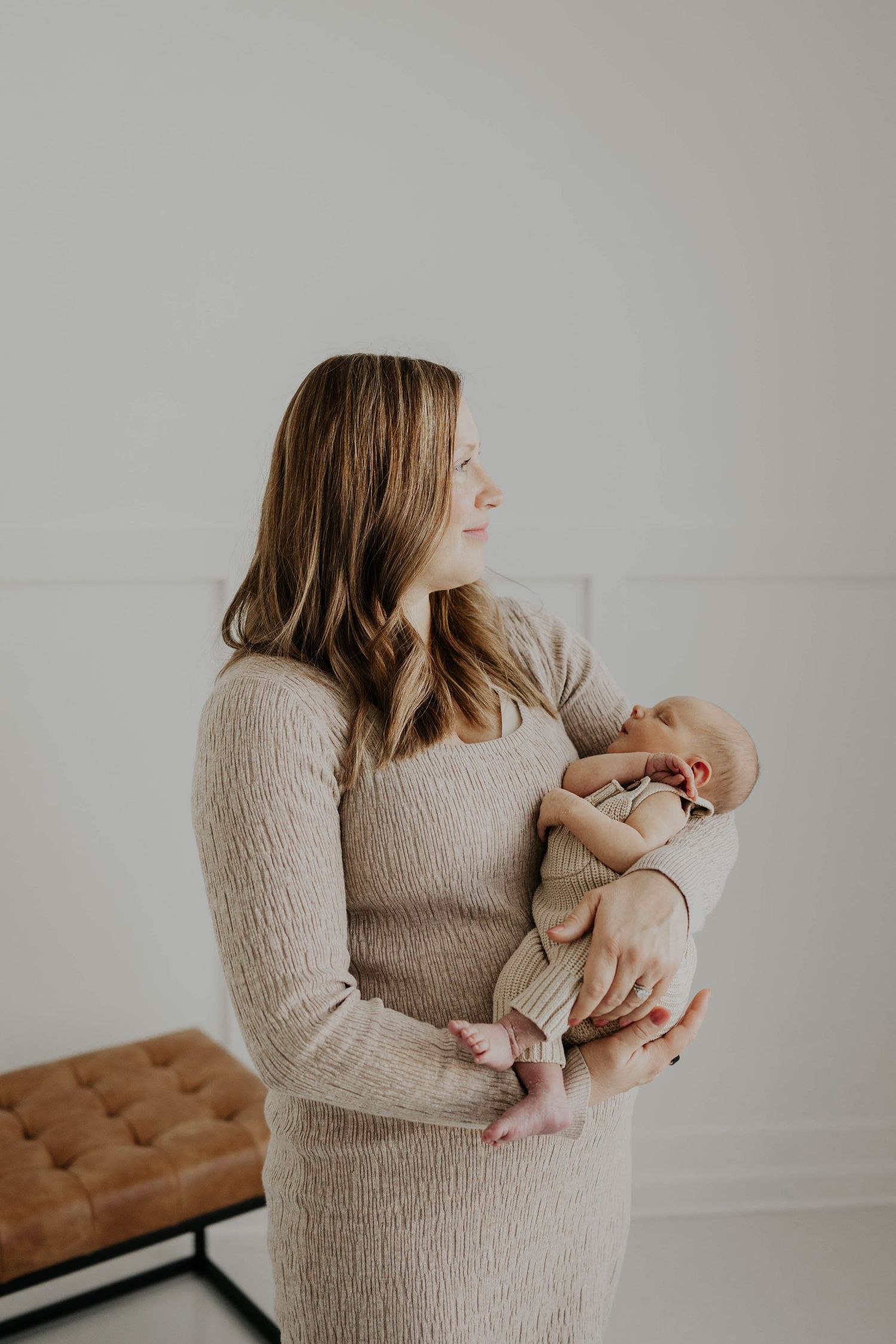 A mom in a beige sweater holds and gazes lovingly at their newborn baby in a minimalist studio setting during newborn photo session in MN.