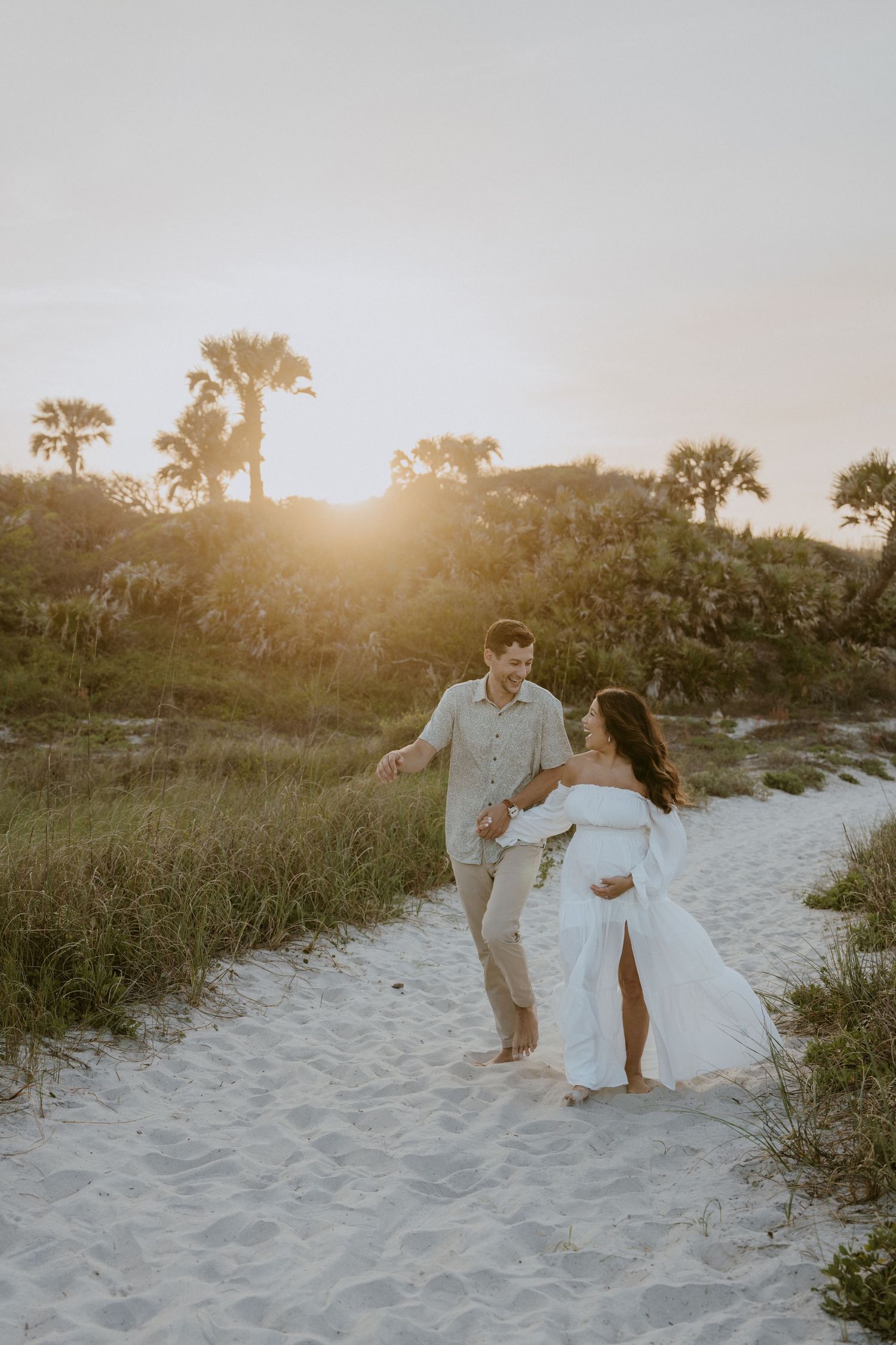A couple in white attire walks along a sandy beach path at sunset with palm trees in the background.