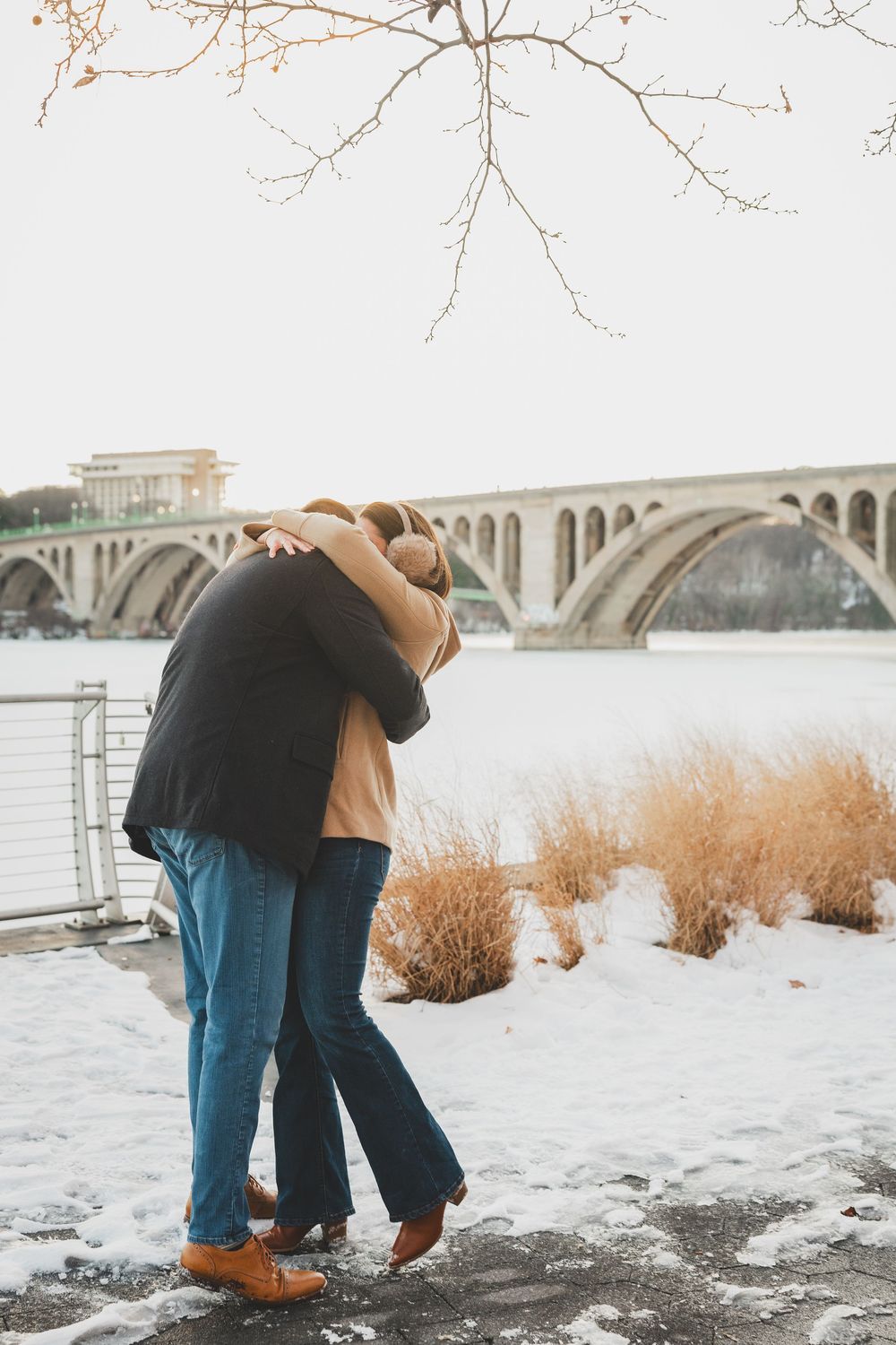 A couple embraces on a snowy riverbank with a bridge visible in the background during winter after popping the question for marriage