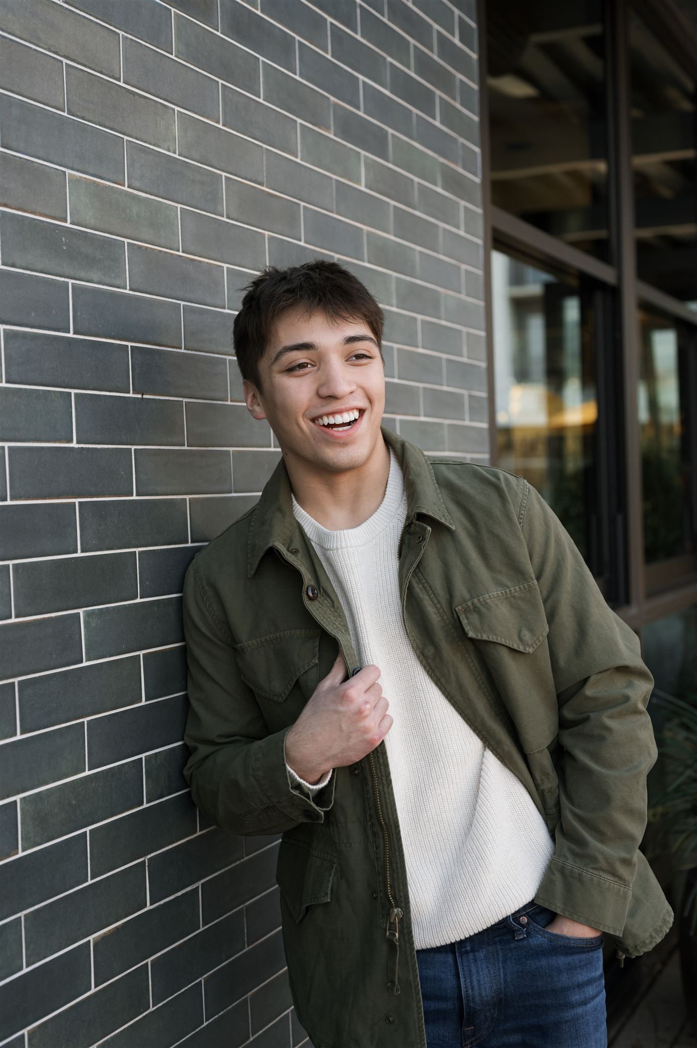 Person wearing olive green jacket and white turtleneck poses against grey brick wall in casual portrait series.