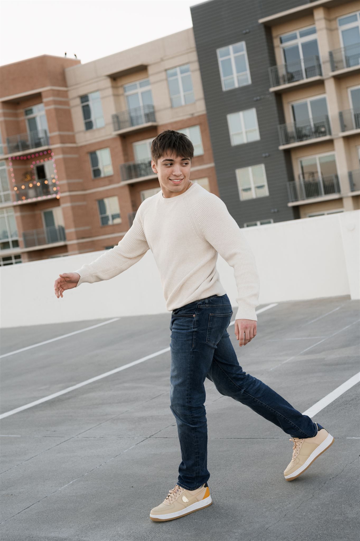 Person in cream sweater and jeans poses on a rooftop parking area with modern apartment buildings in background.