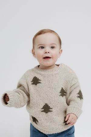 Baby wearing cozy knit sweater with green tree pattern and blue pants against white background.