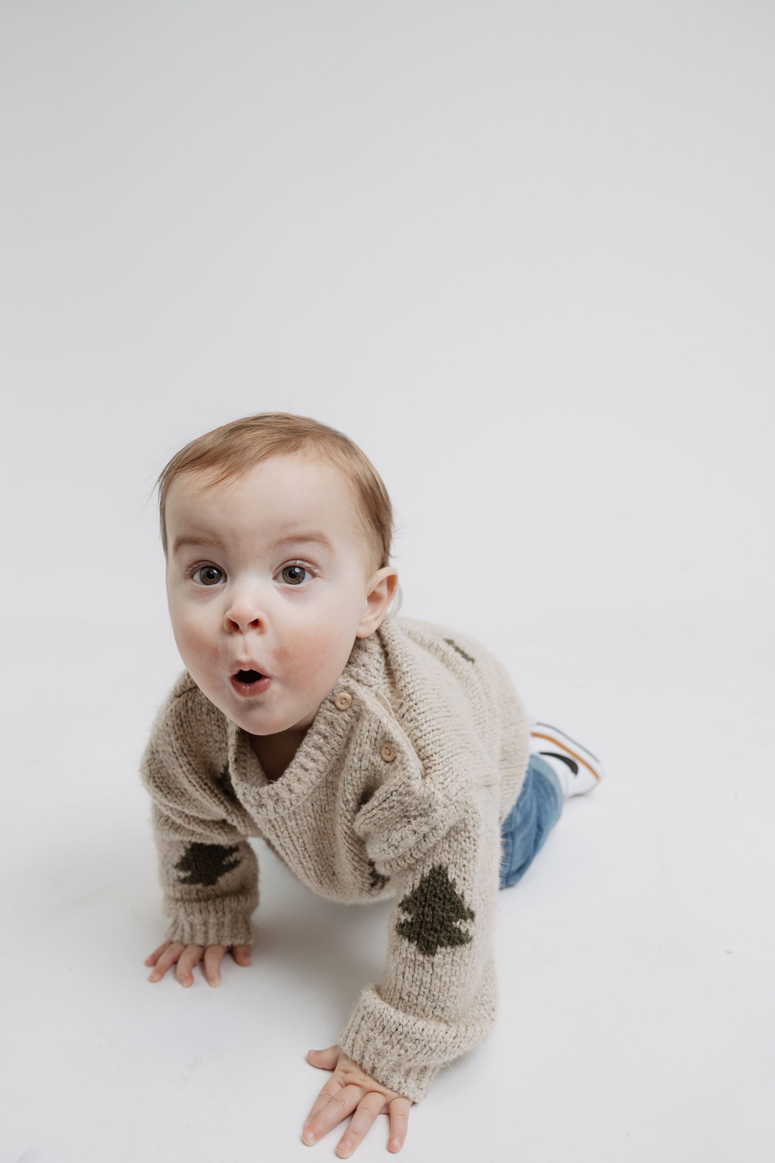 Baby crawling on white background wearing beige sweater with tree designs.