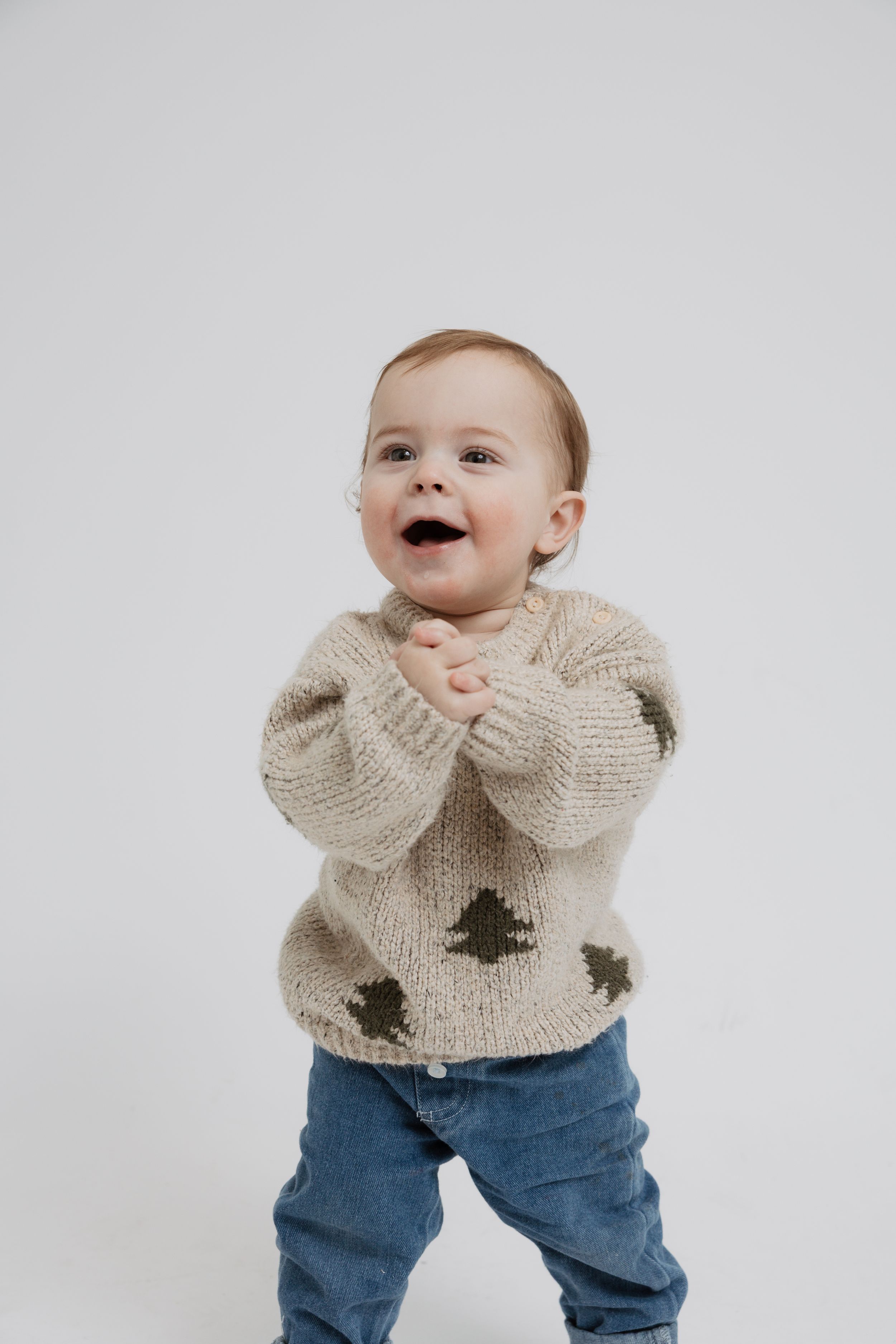 Baby in cream knit sweater with tree designs and blue jeans smiling and making playful expressions against white backdrop.