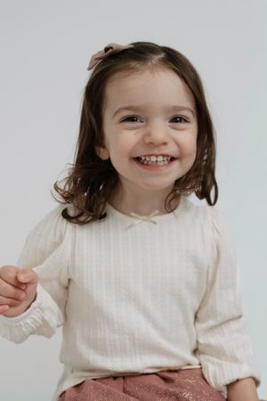 A young child wearing a white sweater and pink pants smiles joyfully against a light background.