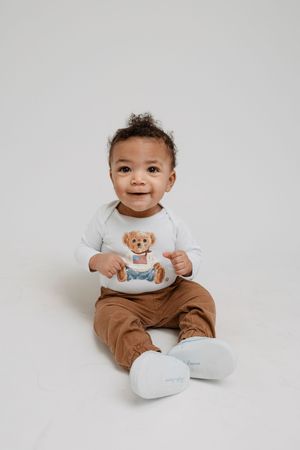 Baby in white long sleeve top and brown pants sits on light gray background showing happy expressions.