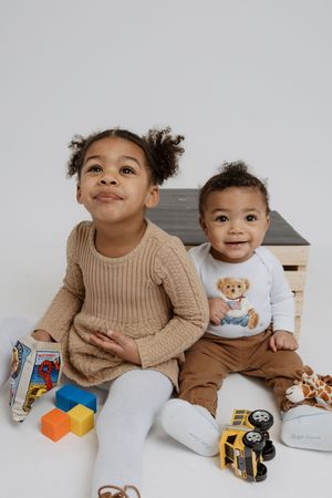 Two children sit together on a white surface wearing cozy sweaters and casual attire.