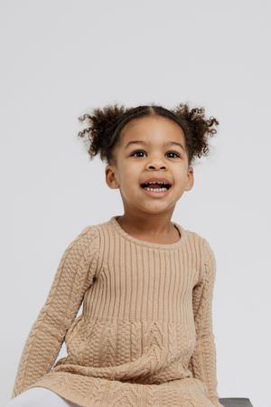 Child wearing a beige cable knit sweater smiles joyfully against a white background.
