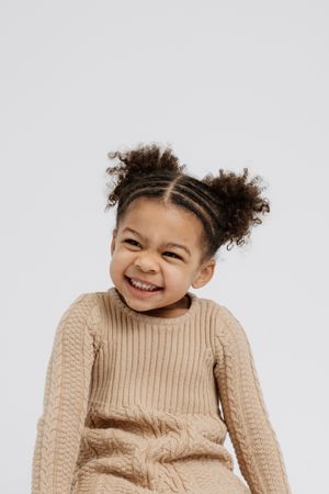 Cheerful child in beige knit sweater with curly hair styled in buns against white background.