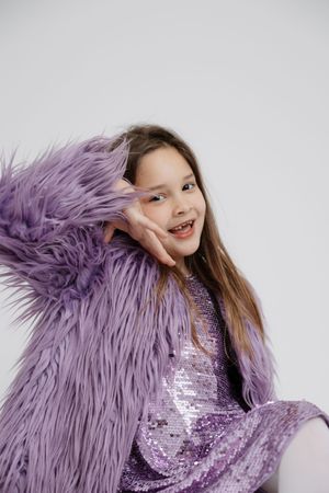 Young model wearing purple sequin dress and fluffy lavender faux fur coat against white background.