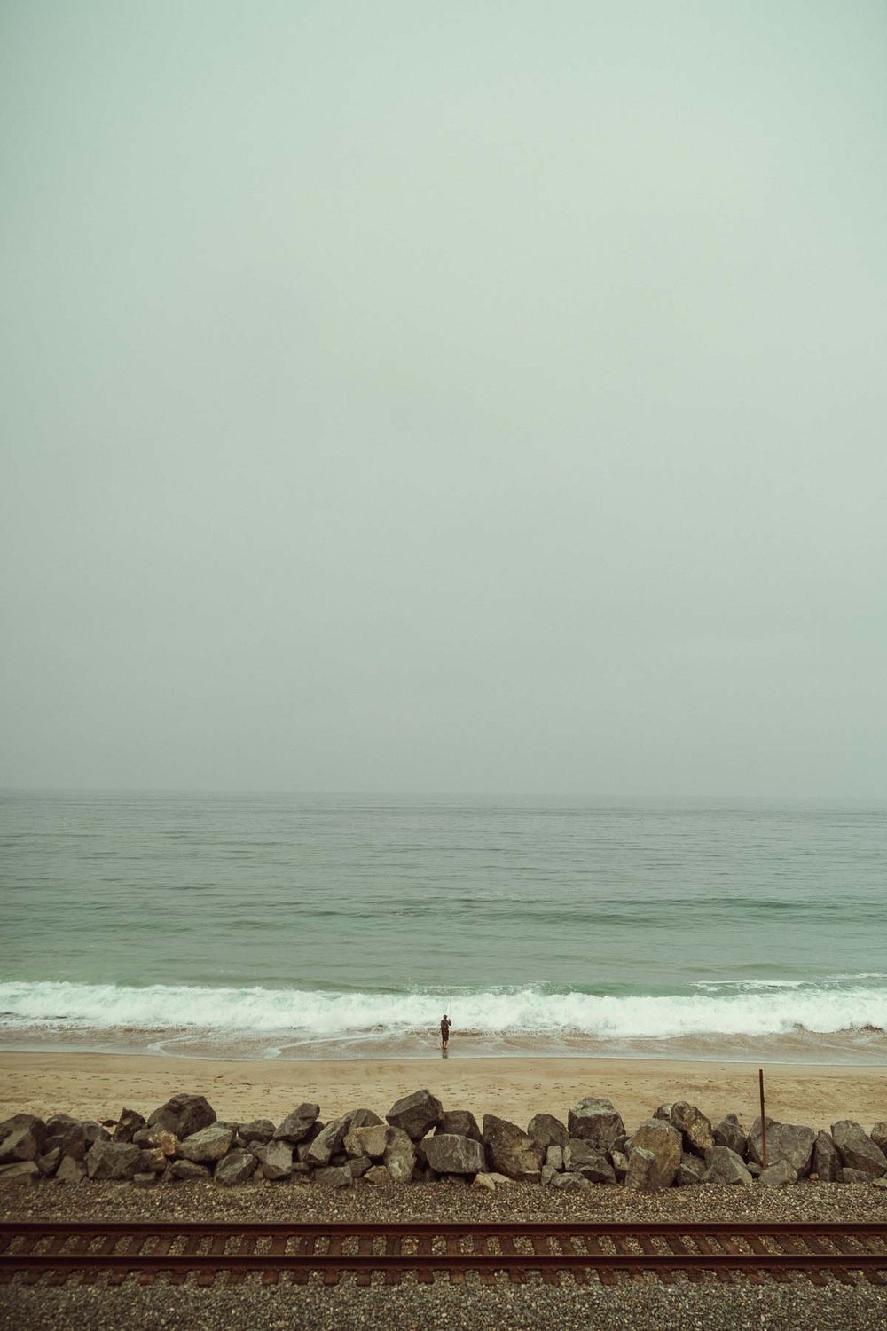 A solitary figure walks along a misty beach with railroad tracks and rocks running parallel to the shoreline.